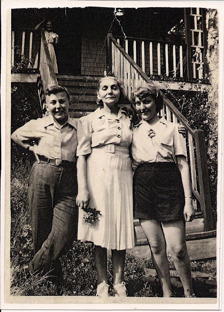 Femme se tenant en bas de l’escalier menant à un chalet, sa fille adolescente à sa gauche et son fils adolescent à sa droite.
