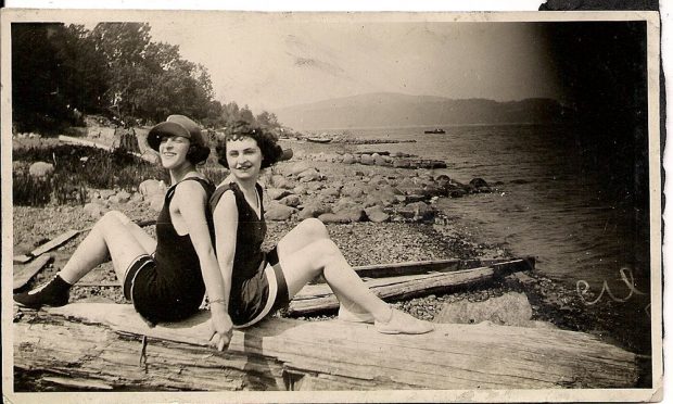 Deux femmes en maillot de bain assises dos à dos sur un gros morceau de bois flotté sur une plage.