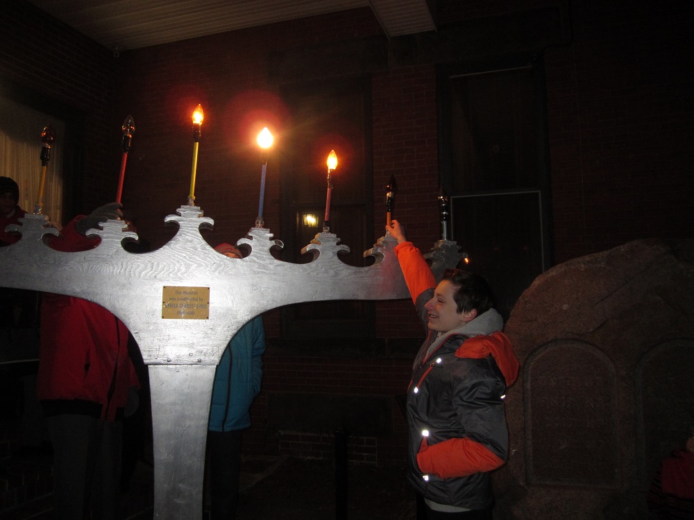 Grande menorah de Hanoukka peinte en gris dont certaines bougies sont allumées. Un adolescent lève le bras pour redresser une bougie.