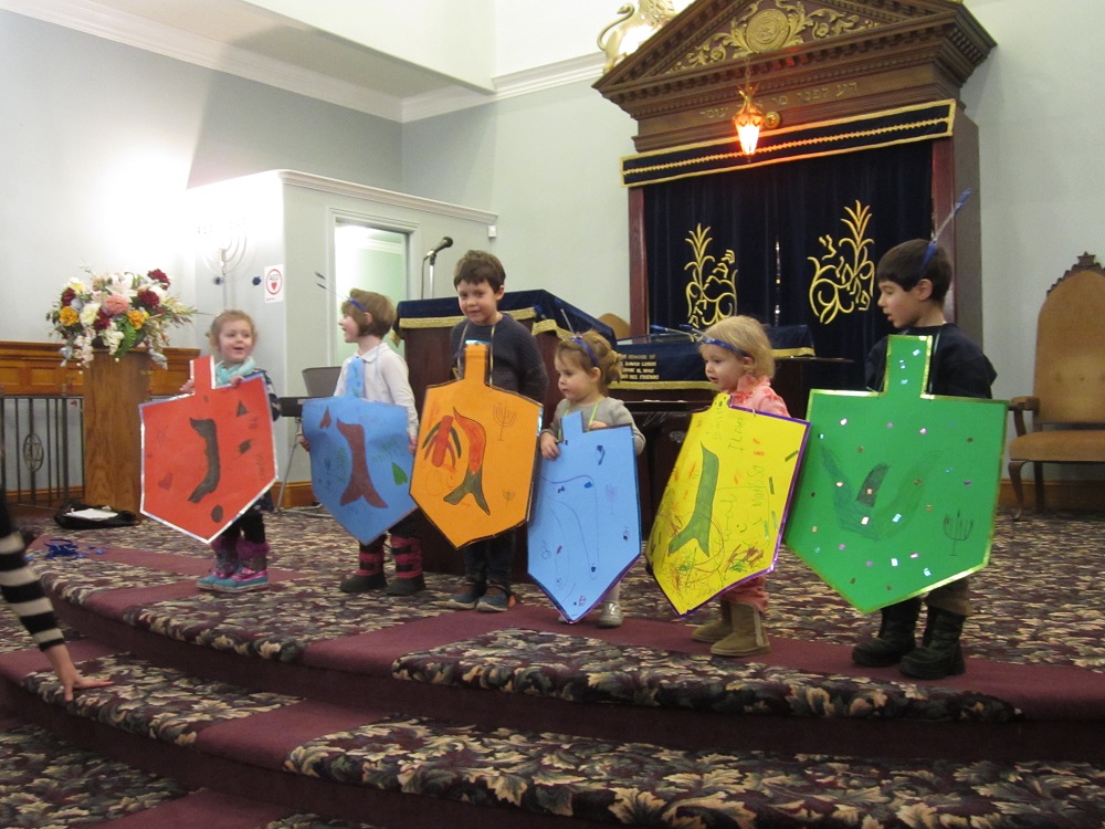 Six enfants sur une estrade devant la synagogue, chacun portant une toupie de Hanoucca en carton grandeur nature.