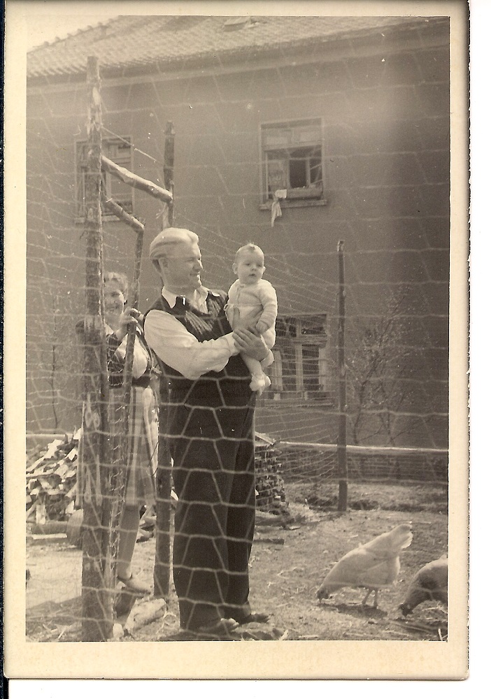 Homme aux cheveux blancs tenant son jeune fils dans les bras tandis que son épouse se trouve à l’arrière-plan. Tous les trois posent derrière une clôture de barbelés.