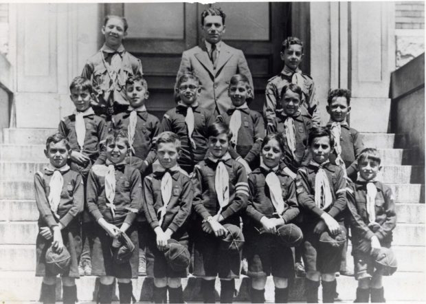 Quatorze garçons en uniforme de louveteau, chapeau à la main, se tenant debout en deux rangées dans l’escalier, leurs dirigeants formant la troisième rangée.