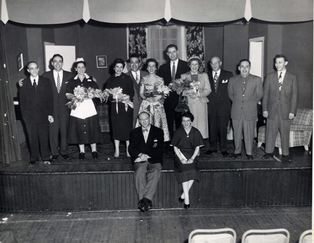 Onze hommes et femmes debout sur la scène en costumes, et un homme et une femme assis devant eux.