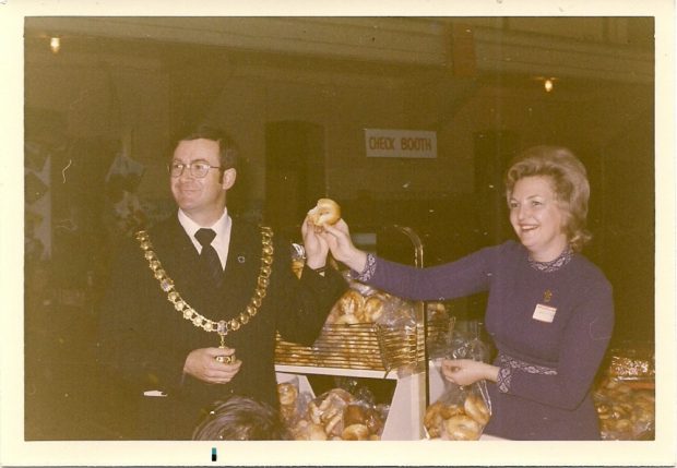 Photographie en couleurs montrant un homme en complet portant la chaîne du maire et une femme devant une table de pâtisseries au cours de la vente de charité de la Hadassah.