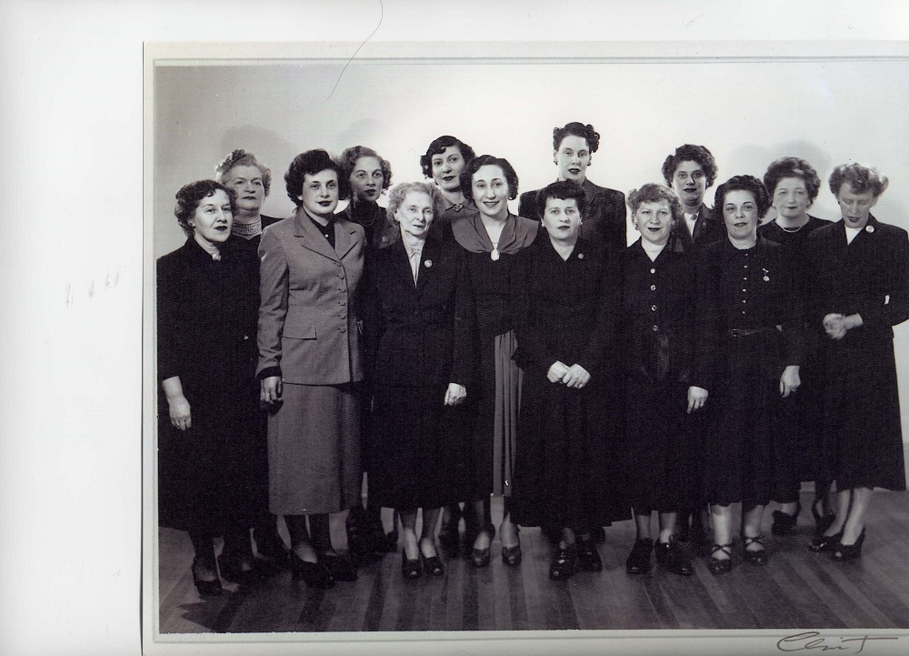 Quatorze femmes d’âge moyen portant une jupe et une veste se tenant debout en deux rangées.