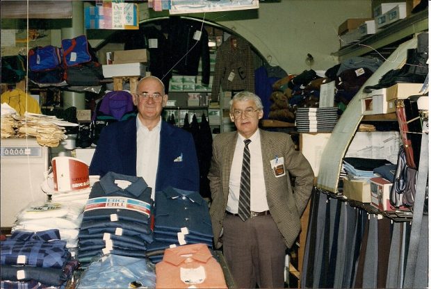 Deux hommes en complet derrière un comptoir exposant des chemises de travail pour homme et, sur la droite, des rayons bien garnis.