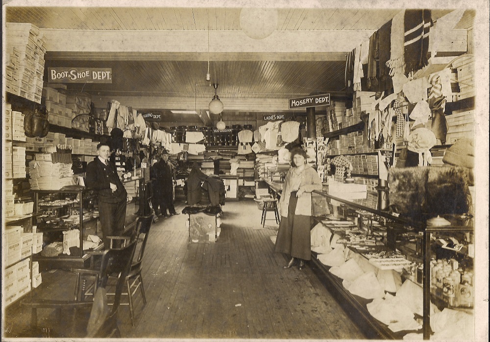 Vue à l’intérieur d’un étroit magasin d’articles de mercerie dont les comptoirs et rayons sont remplis de boîtes, d’un côté comme de l’autre. Un homme se tient à gauche et une femme à droite.
