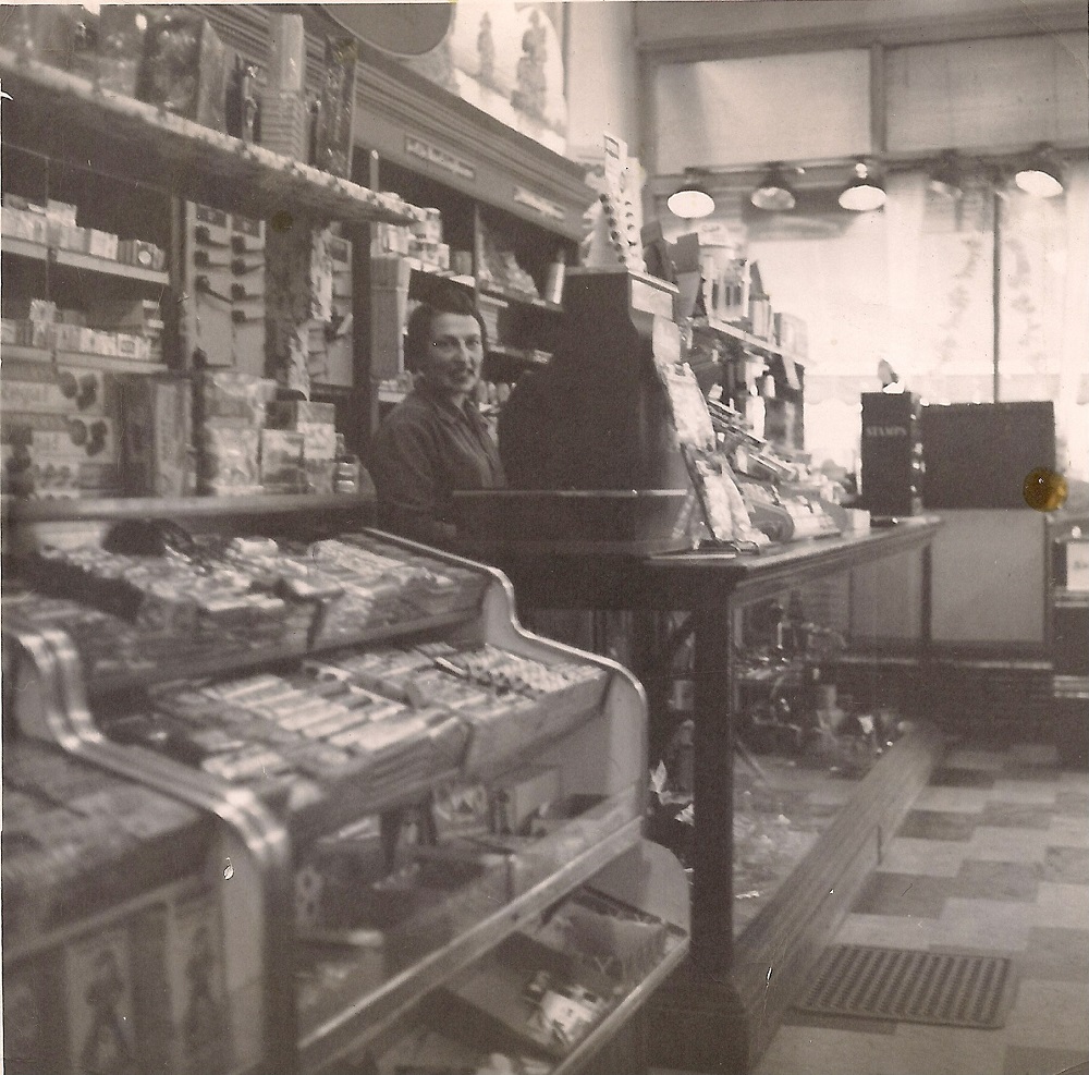 Photographie montrant l’intérieur d’un magasin dont le comptoir regorge de petits articles et dont les rayons sont remplis de boîtes. Une jeune femme se tient derrière une grosse caisse enregistreuse posée sur le comptoir.