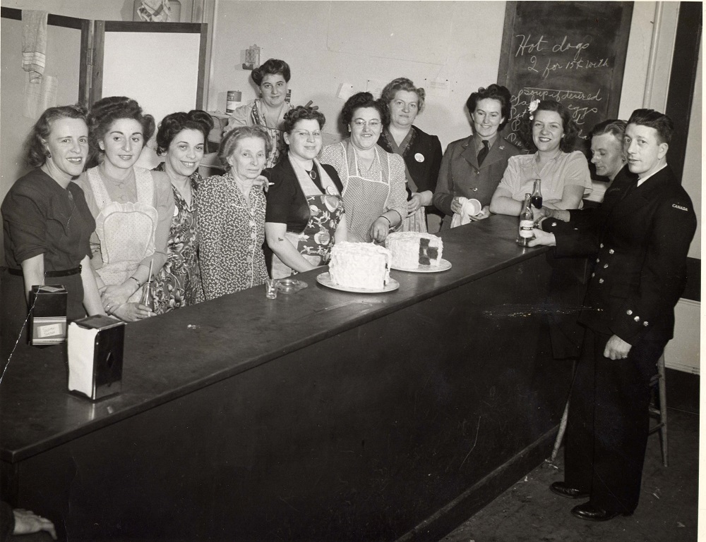 Dix femmes alignées derrière un long comptoir, certaines portant un tablier, et deux hommes portant des uniformes de la marine au bout du comptoir.