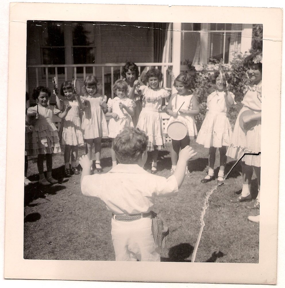 Garçon debout dos à l’appareil photo dirigeant un groupe de filles en robes d’été jouant de divers instruments, notamment du triangle et du tambourin.