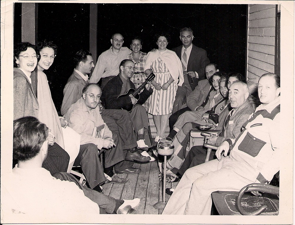 Seize hommes et femmes assis sur une véranda en bois. Un des hommes tient un ukulélé.