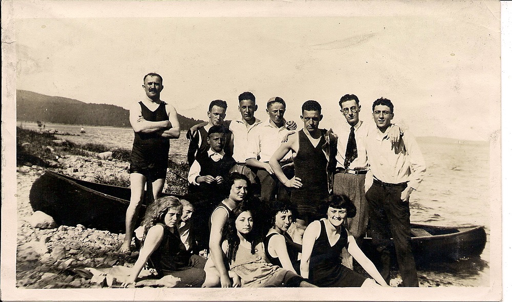 Treize amis, dont certains en maillot de bain et d’autres en pantalon et chemise rassemblés devant un canot sur une plage.