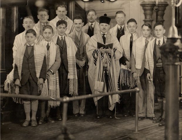 Treize hommes et garçons en complet-veston portant des châles de prière alors que le rabbin tient un livre de prières ouvert.