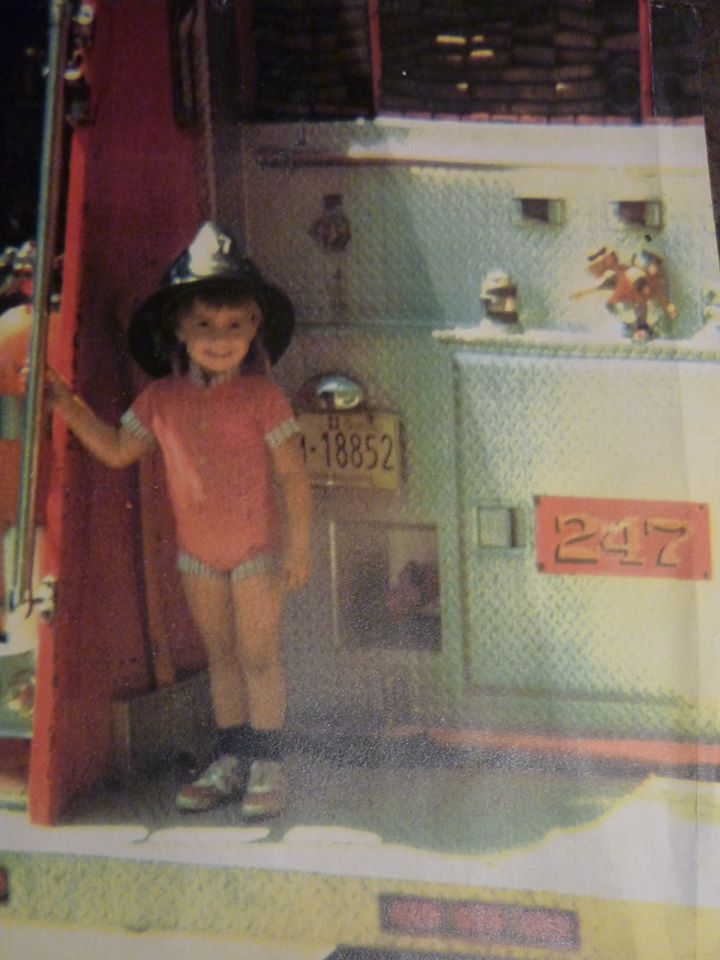 Photographie en couleurs d’une jeune fille d’environ quatre ans debout à l’arrière d’un camion de pompier, souriante et portant sur sa tête un casque de pompier. 