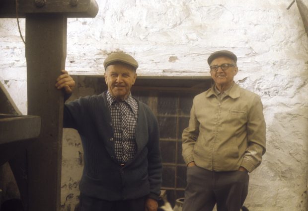 Photographie de deux hommes septuagénaires tout sourire devant un mur de béton blanc. 