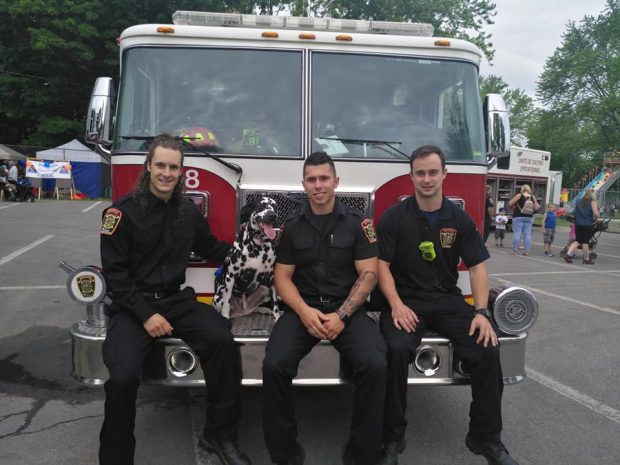Photographie de trois hommes en uniforme civil de pompier assis sur le devant d’un camion de pompier avec un chien dalmatien.
