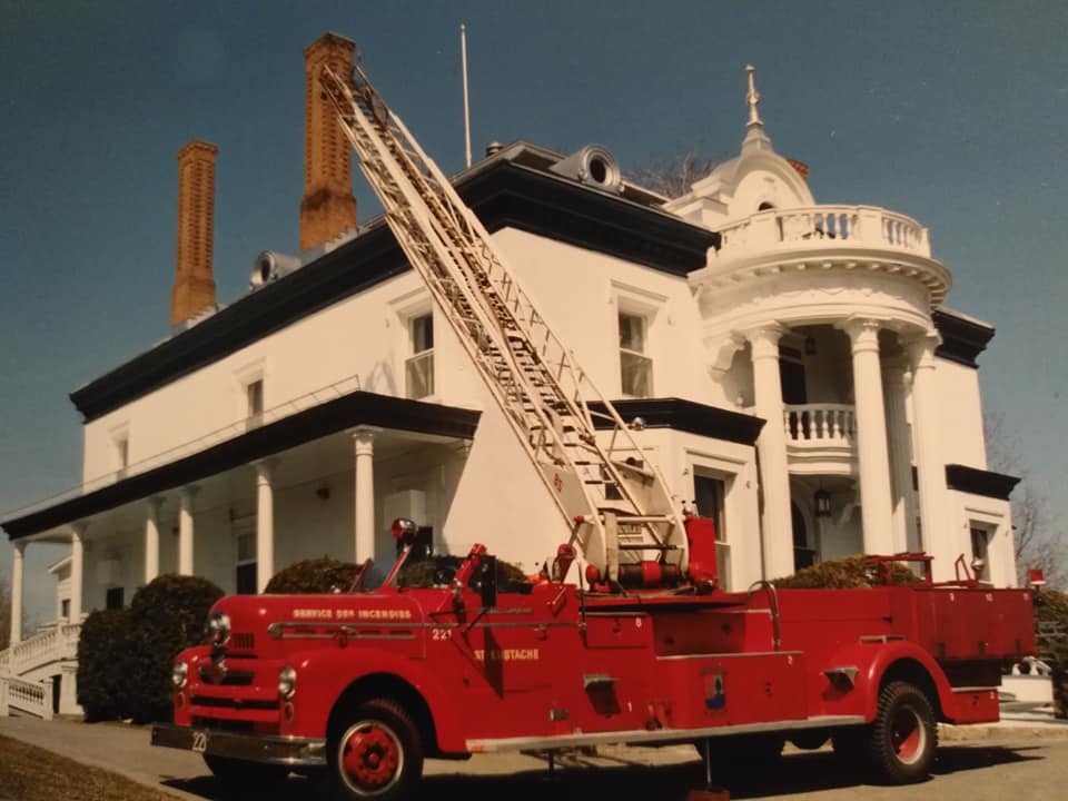 Photographie d’un camion de pompier rouge et convertible hissant son échelle aérienne devant le présentoir d’un manoir blanc datant du XIXe siècle.