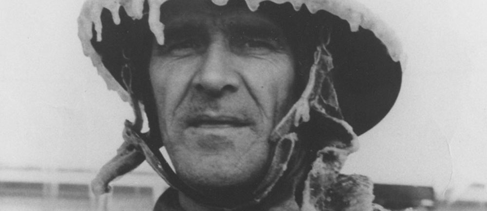 Photographie en noir et blanc d’un homme au visage paisible, arborant un uniforme de pompier couvert de glace. 