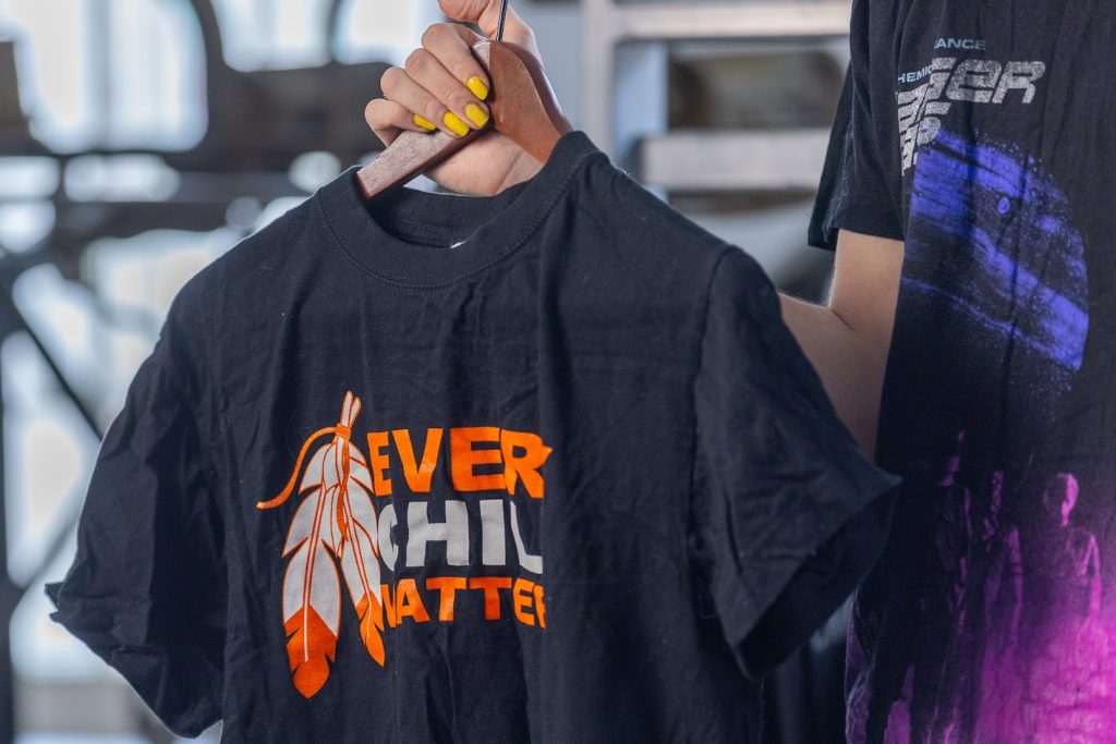 Un t-shirt noir sur un cintre en bois. Un texte orange et blanc se lit EVERY CHILD MATTERS (chaque enfant compte). À gauche du texte, il y a un motif : deux plumes, en orange et blanc.