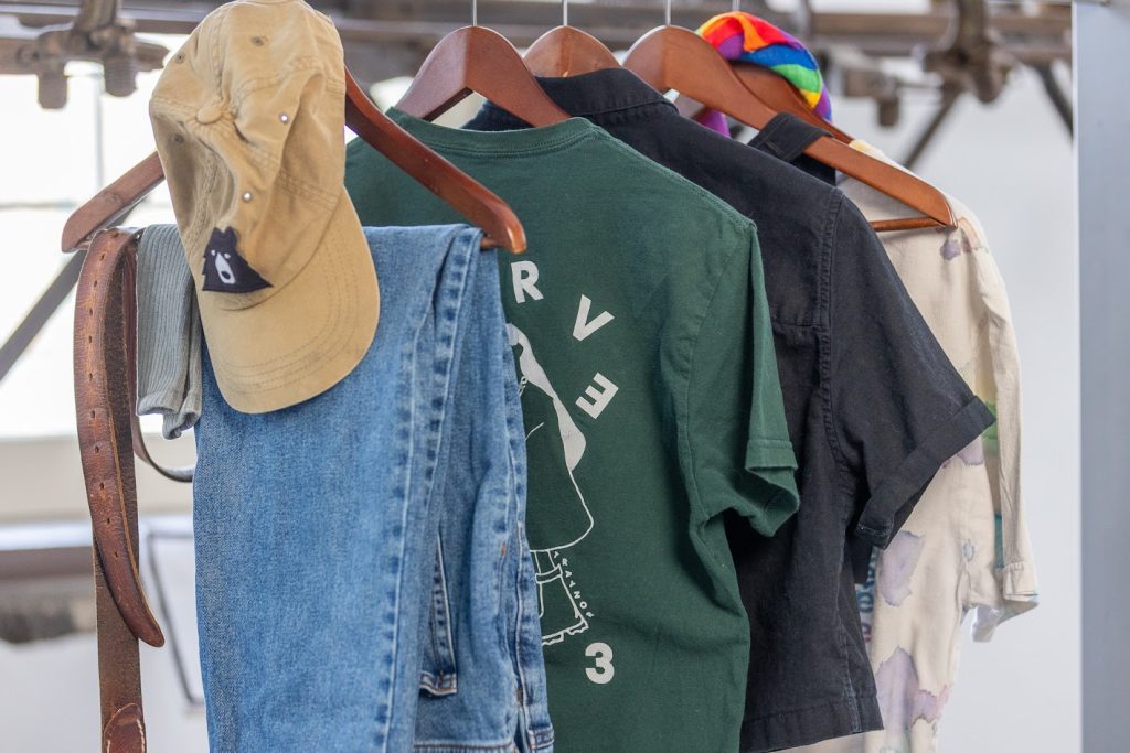 Des vêtements suspendus à cinq cintres en bois, surtout de couleurs neutres, à l’exception d’un mouchoir à rayures arc-en-ciel noué au haut d’un cintre.