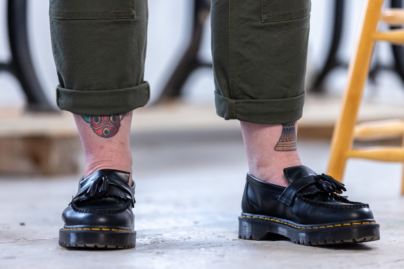 Gros plan de jambes, à partir du mollet. La personne porte des mocassins Dr Martens et un pantalon vert foncé à revers. Des tatouages multicolores sont visibles sur ses jambes. 