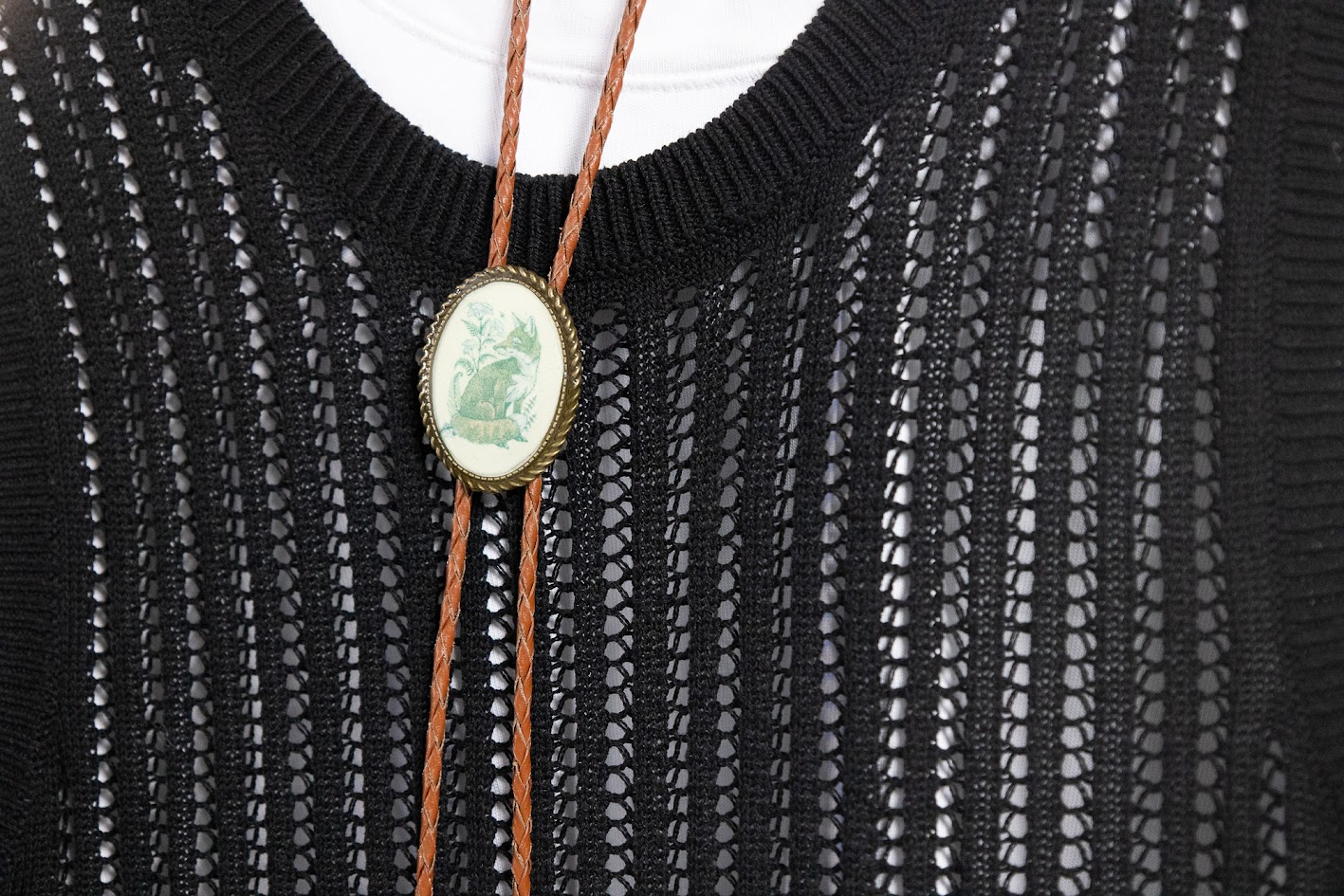 Une cravate western en cuir marron clair, maintenue par un médaillon ovale orné d’un dessin de renard, en vert. La cravate est portée avec un haut noir en tricot sur un t-shirt blanc.
