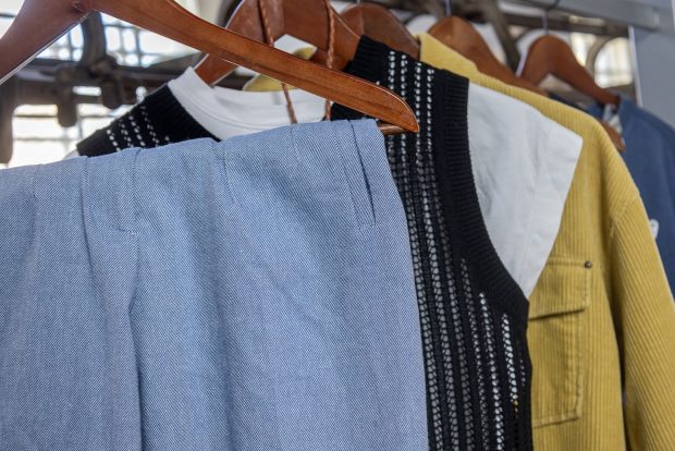 Des vêtements sur des cintres en bois, suspendus à un support. La gamme de vêtements, de couleurs et styles variés, comprend un pantalon en sergé bleu et blanc, un gilet noir en tricot et une veste en velours côtelé jaune.