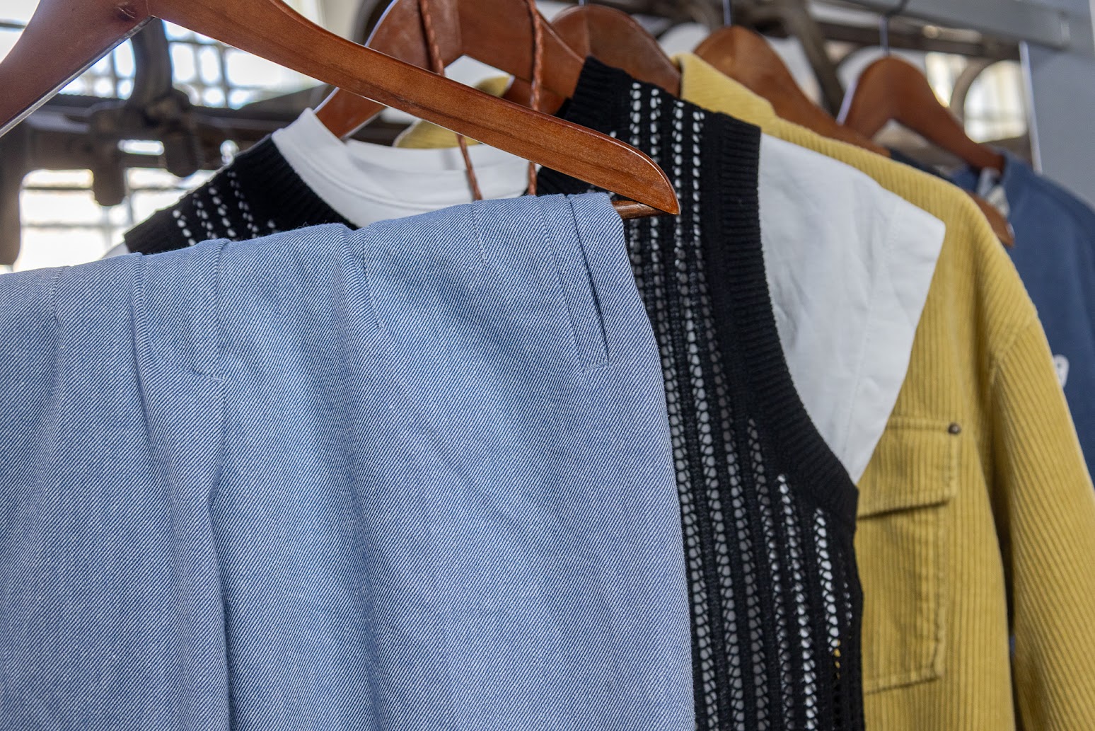 Des vêtements sur des cintres en bois, suspendus à un support. La gamme de vêtements, de couleurs et styles variés, comprend un pantalon en sergé bleu et blanc, un gilet noir en tricot et une veste en velours côtelé jaune.