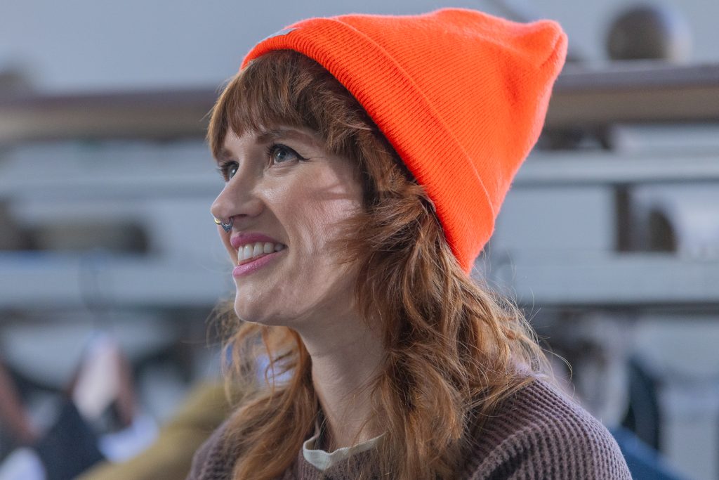 Portrait d’une personne inclinée vers la gauche, souriant et levant les yeux. Iel porte un chapeau en tricot orange vif.