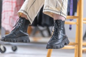 Photo des pieds d’une personne assise sur un banc. Iel porte des bottes noires à lacets et le bas de son pantalon est visible. Le pantalon blanc cassé est illustré de détails en noir lui donnant un effet 2D.