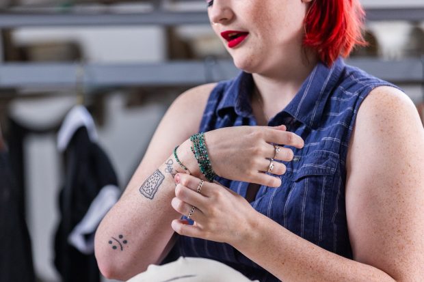 Une personne levant un bras et montrant un tatouage manuscrit. Iel a des bracelets de perles et des bagues en argent, et deux autres tatouages sur les bras.