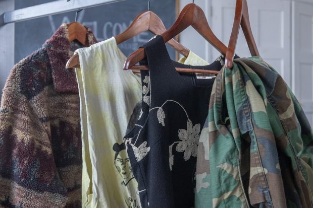 Quatre vêtements alignés, suspendus à des cintres en bois, montrant une gamme de styles. Il y a un cardigan en laine polaire à motif paysage, un débardeur orné d’un motif noir, une robe noire sans manches avec un motif floral blanc et un pantalon camouflage.