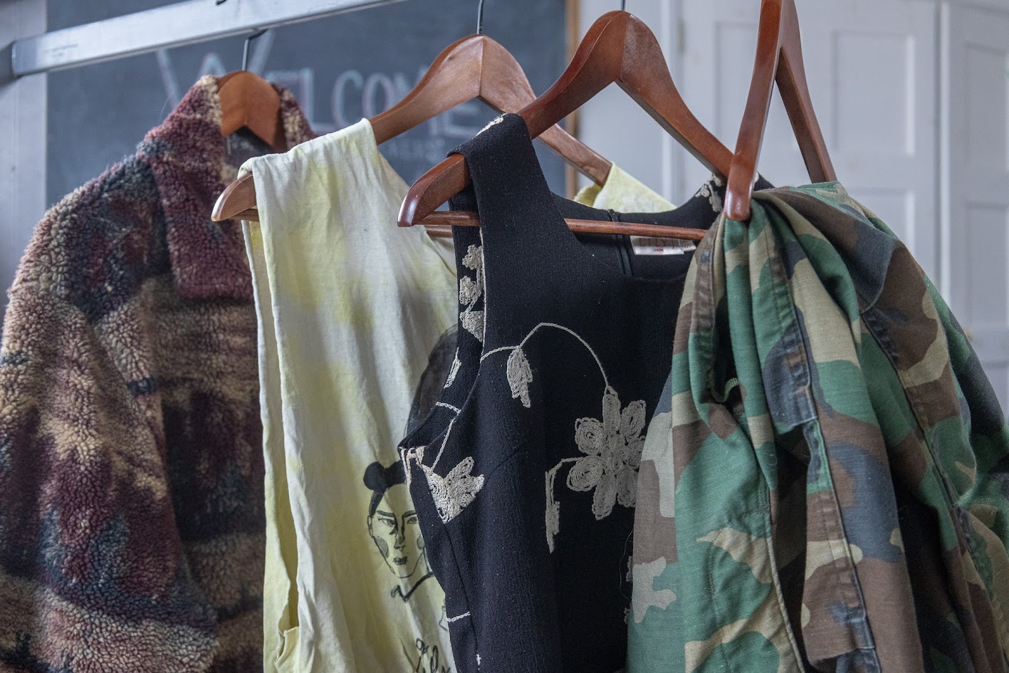 Quatre vêtements alignés, suspendus à des cintres en bois, montrant une gamme de styles. Il y a un cardigan en laine polaire à motif paysage, un débardeur orné d’un motif noir, une robe noire sans manches avec un motif floral blanc et un pantalon camouflage. 