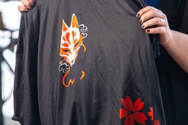 Des mains tiennent un vêtement noir orné d’un visage kitsune ou esprit renard, en blanc, orange et rouge et de fleurs rouges.