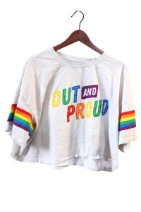 Un haut court blanc cassé sur un cintre en bois. Le texte aux couleurs de l’arc-en-ciel se lit « OUT AND PROUD » – assumée et fière. Il y a des rayures arc-en-ciel sur les manches.
