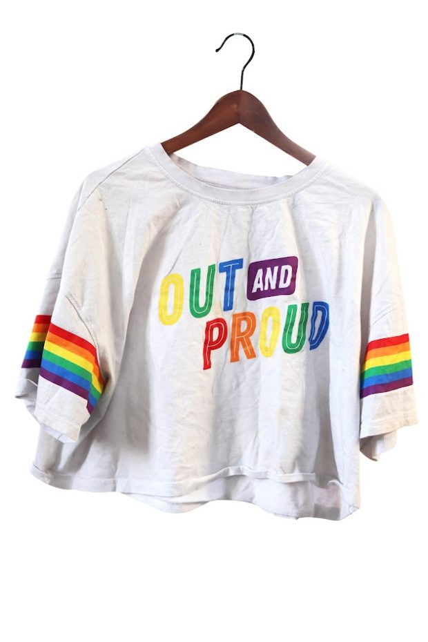 Un haut court blanc cassé sur un cintre en bois. Le texte aux couleurs de l’arc-en-ciel se lit « OUT AND PROUD » – assumée et fière. Il y a des rayures arc-en-ciel sur les manches.