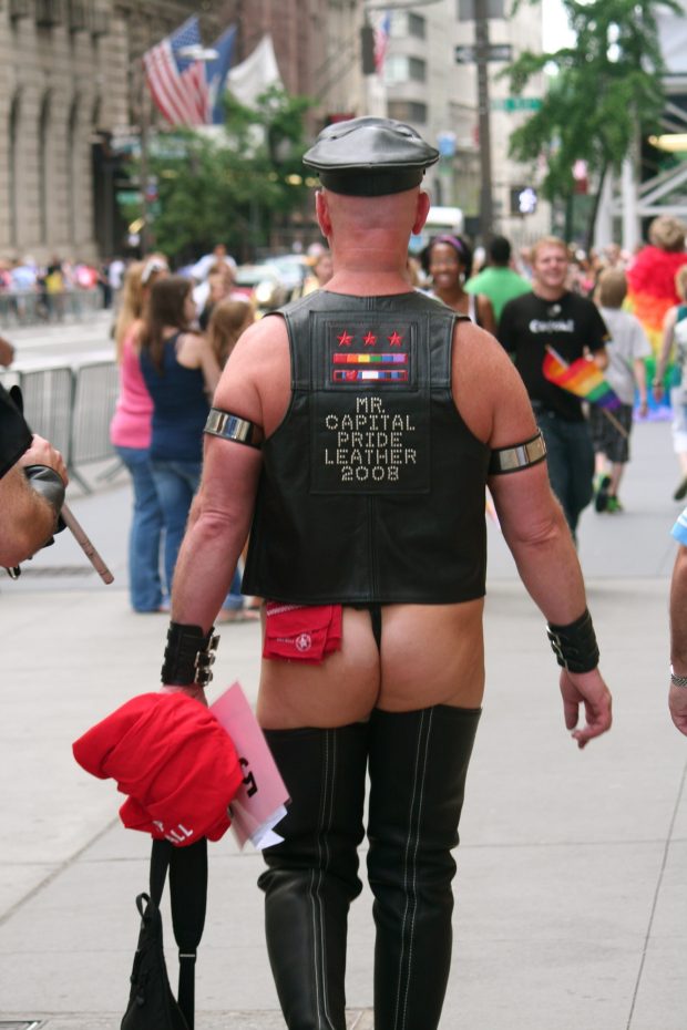 Un homme vu de dos marche sur un trottoir achalandé. Il porte des bottes cuissardes en cuir, un string et un mouchoir rouge à la ceinture. Il porte aussi une casquette en cuir et un gilet en cuir orné de l’inscription MR CAPITAL PRIDE LEATHER 2008, en lettres scintillantes.