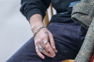 Une main portant des anneaux et un bracelet chaine en argent reposant sur la jambe d’une personne portant un jean pourpre.