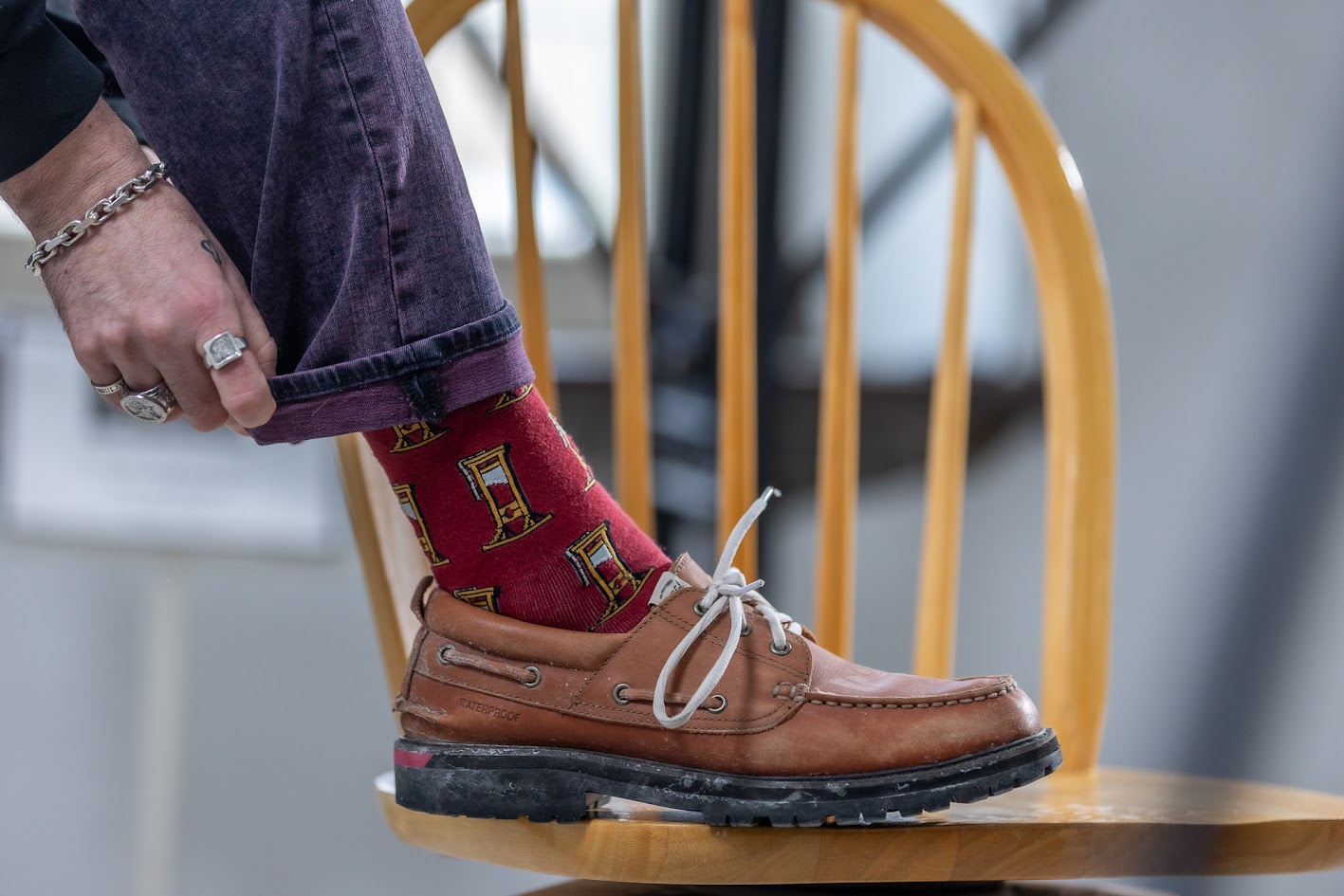 Un pied repose sur une chaise en bois. La personne porte des chaussures bateau marron et un jean pourpre roulé pour montrer ses chaussettes rouges au motif de guillotines. La main qui roule le bas du pantalon porte des bagues et un bracelet en argent. 
