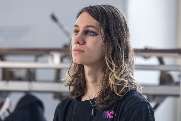 Portrait en plan serré, à partir des épaules. La personne est inclinée vers la gauche et regarde droit devant elle. Elle porte un t-shirt noir avec un petit texte « MCR » violet sur le côté droit. Elle porte du maquillage pour les yeux violet et jaune.