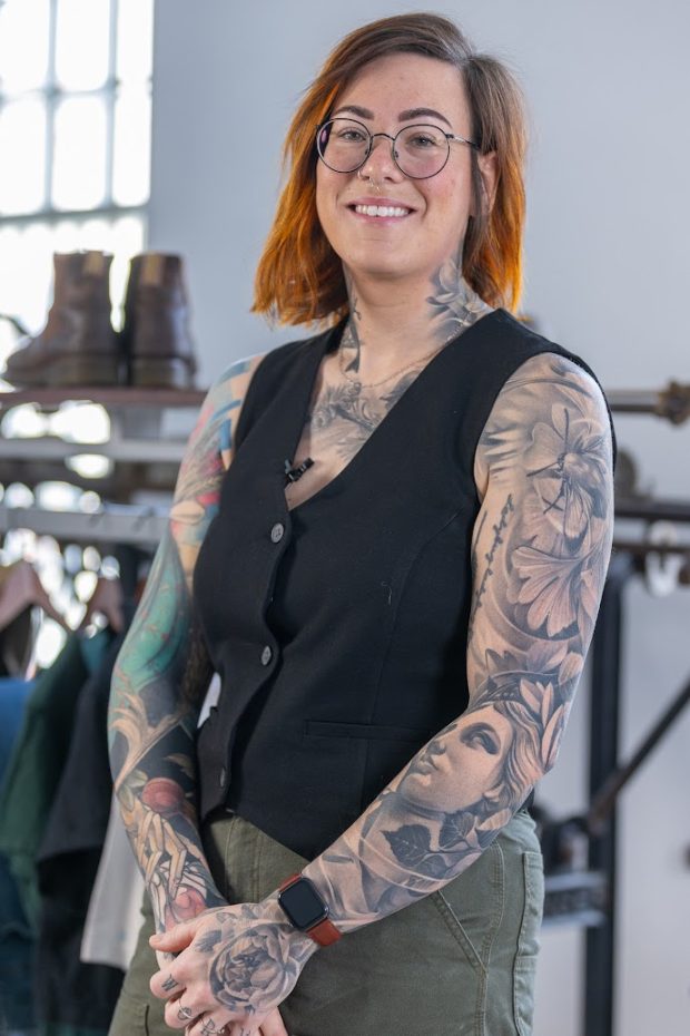 Portrait en plan américain. La personne sourit, porte des lunettes rondes à monture noire et regarde la caméra. Elle porte un pantalon vert et une veste sans manches boutonnée. Des tatouages couvrent ses bras et sa poitrine et montent le long de son cou.