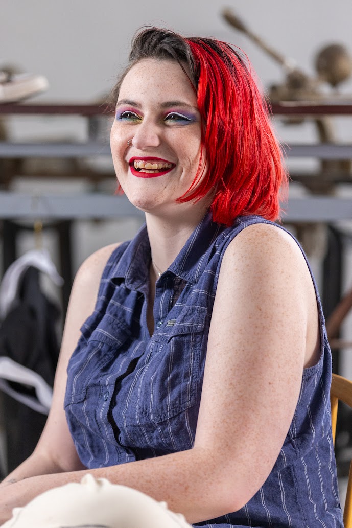 Portrait en plan taille. La personne est inclinée vers la gauche et regarde droit devant elle en riant. Iel a les cheveux rouge vif et le maquillage des yeux est violet, rose et blanc. Iel porte une chemise boutonnée, sans manches, bleu foncé à fines rayures blanches. 