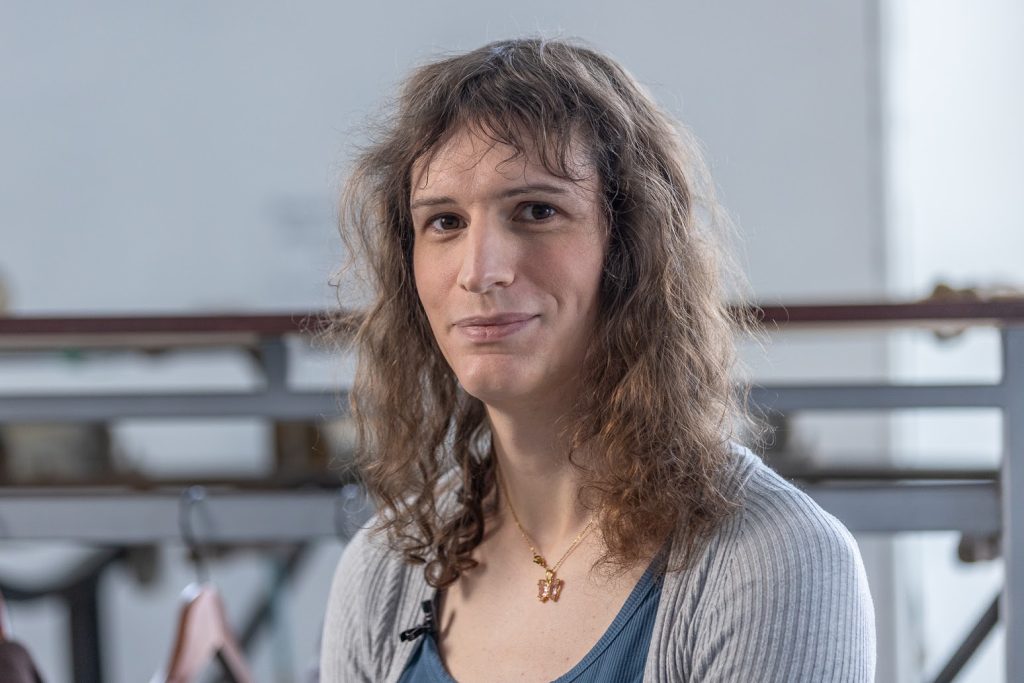 Portait en buste. La personne sourit et regarde la caméra. Elle porte un cardigan gris côtelé et une chemise bleu foncé à encolure dégagée, ainsi qu’un collier doré.