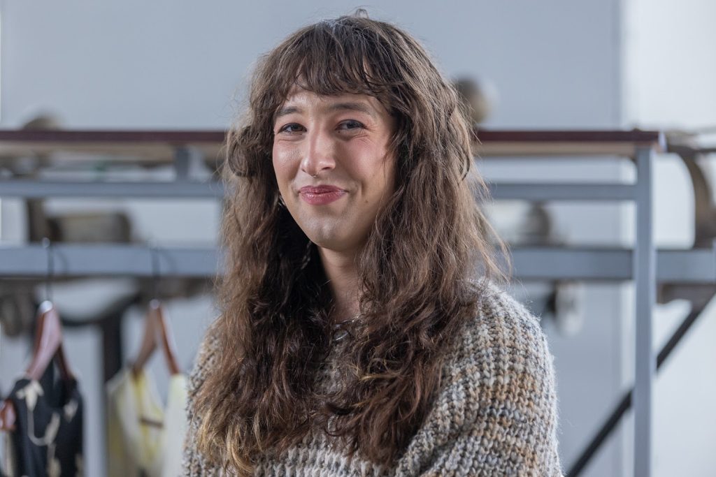 Portrait en buste. La personne sourit et regarde la caméra. Elle porte un pull en tricot gris, brun et orange.