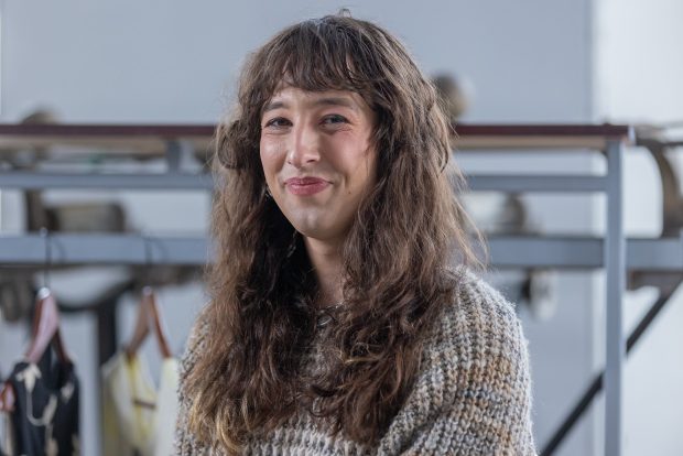 Portrait en buste. La personne sourit et regarde la caméra. Elle porte un pull en tricot gris, brun et orange.