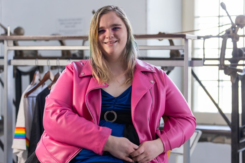Portrait en plan américain. La personne sourit et regarde la caméra. Elle porte une veste en simili cuir rose sur une chemise bleu foncé et noir avec une boucle argentée. Ses yeux sont maquillés en rose.
