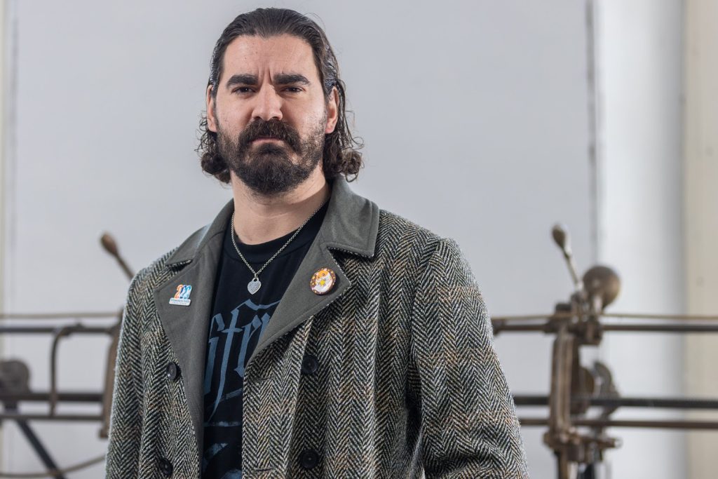 Portrait en plan taille. Un barbu regarde dans la caméra d’un air sérieux. Il porte un caban gris et noir avec une épinglette sur chaque revers. En dessous, il porte une chemise noire avec du lettrage bleu.