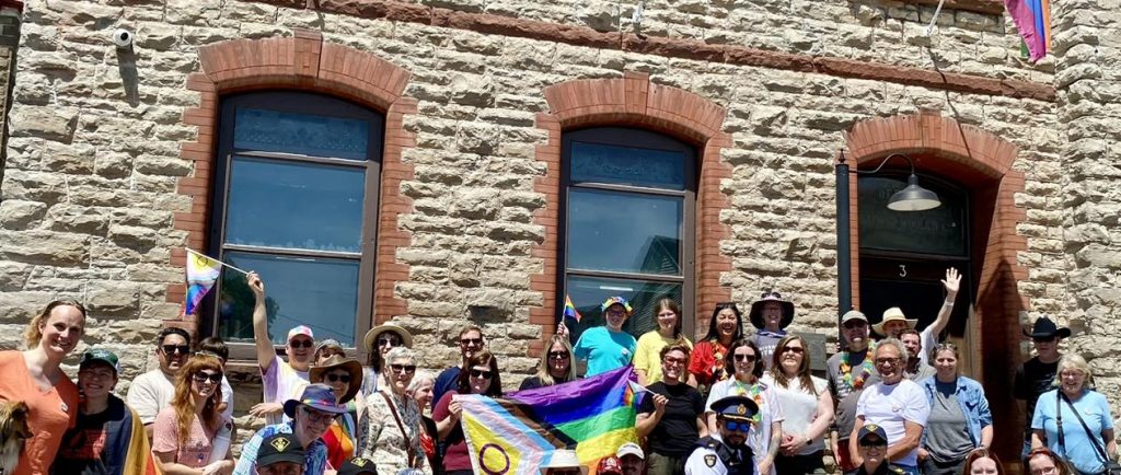 Un groupe de personnes est rassemblé à l’extérieur d’un bâtiment en pierre, en place pour prendre une photo. Un drapeau de la Fierté est brandi au milieu de la foule, et un autre flotte sur un mât au-dessus de l’entrée du bâtiment. Les gens portent eux-mêmes de plus petits drapeaux de la Fierté. Ils sourient à la caméra.
