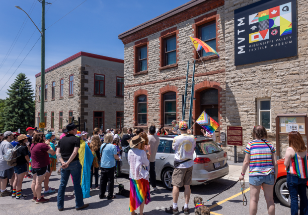 Une foule est rassemblée devant un bâtiment en pierre. Une échelle est déployée au-dessus de l’entrée du bâtiment. Un drapeau de la Fierté flotte au mat.