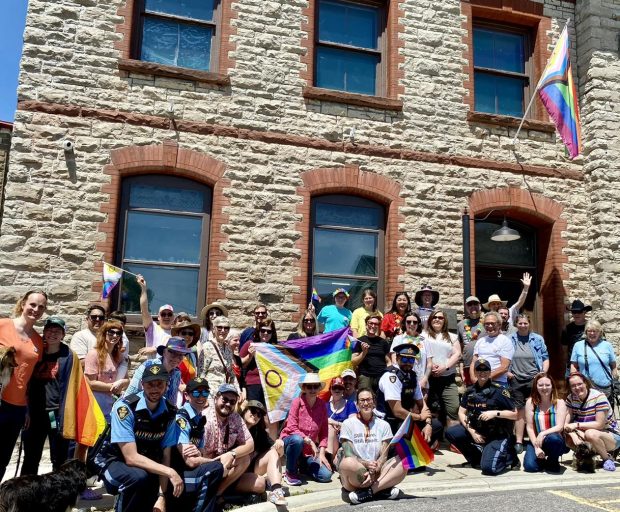 Un groupe de personnes est rassemblé à l’extérieur d’un bâtiment en pierre, en place pour prendre une photo. Un drapeau de la Fierté est brandi au milieu de la foule, et un autre flotte sur un mât au-dessus de l’entrée du bâtiment. Les gens portent eux-mêmes de plus petits drapeaux de la Fierté. Ils sourient à la caméra.